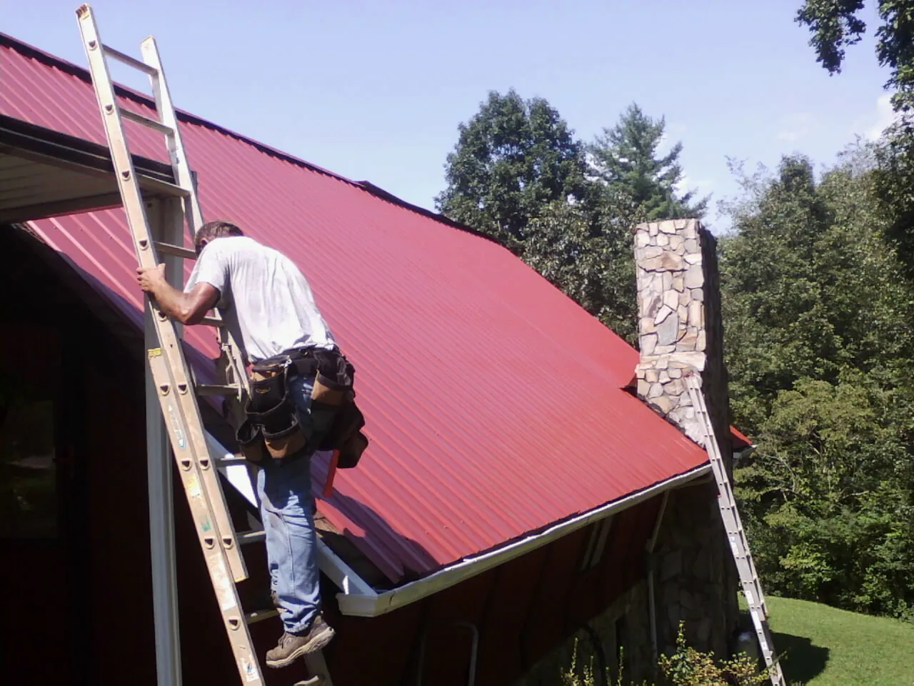 Roofer on a ladder beside a red metal roof and stone chimney — Soffit & Fascia Repair by Roofing Brothers in Mayesville